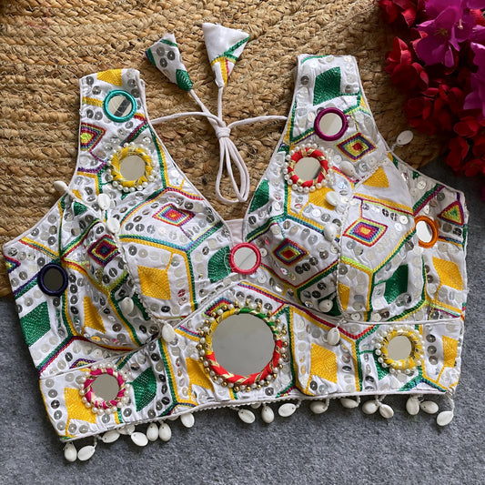 Traditional white Gujarati-style blouse featuring vibrant multicolor geometric embroidery, large circular mirrors framed with colorful beads, intricate shell accents along the hem, and tasseled tie-back detail, displayed flat on a grey carpet next to a woven mat and fuchsia artificial flowers.