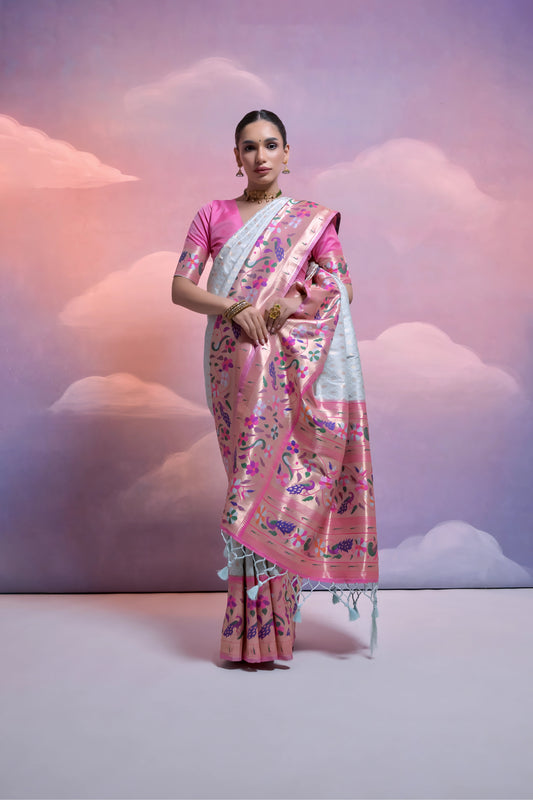 Woman wearing a white Banarasi silk saree with a pink floral zari border and pallu, paired with a pink blouse and traditional jewelry, posing in a pastel-themed studio with dreamy cloud backgrounds.
