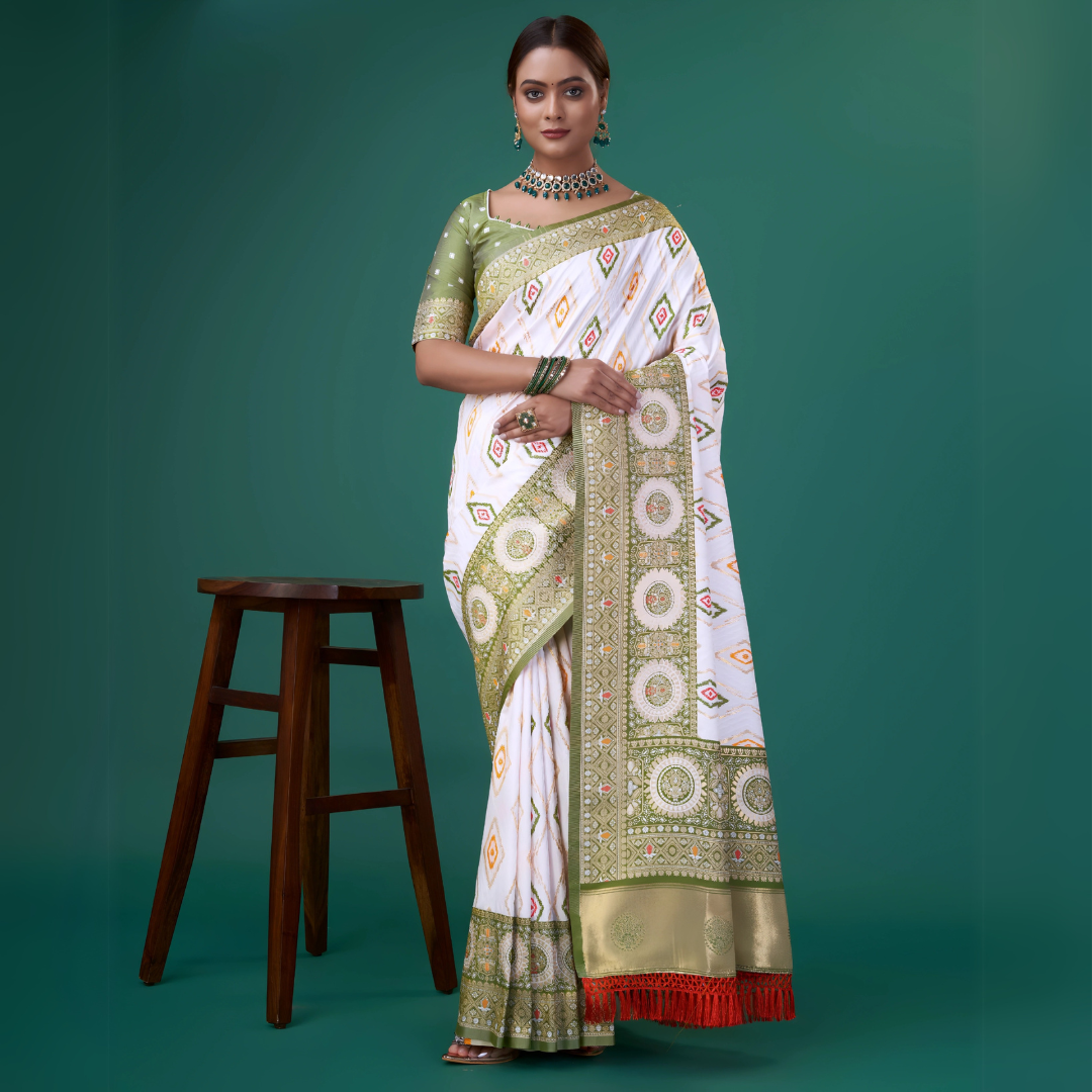 Woman wearing a white silk saree with intricate green and gold border, featuring circular and diamond-shaped geometric motifs, paired with a green polka dot blouse and traditional jewelry, standing beside a wooden stool in a studio with a green backdrop.