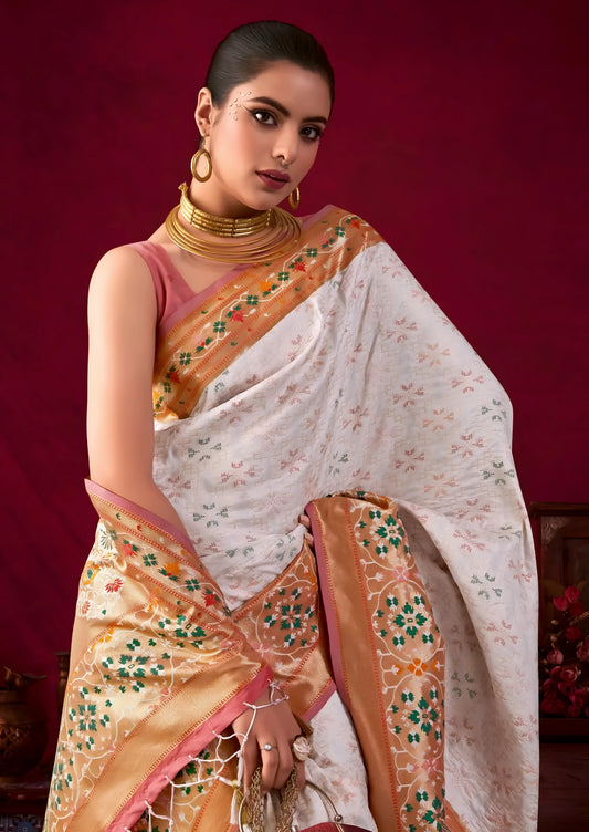 Woman wearing a white silk saree with an orange and gold floral embroidered border, accessorized with gold jewelry and a maroon handbag, standing in front of a vintage wooden table and decor against a deep red backdrop.