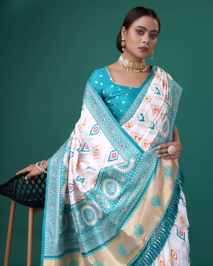 Woman wearing a white silk saree with turquoise blue border and intricate circular and diamond-shaped geometric motifs, paired with a matching turquoise blouse adorned with polka dots, accessorized with traditional jewelry, posing beside a modern black chair in a studio with a green backdrop.