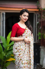 Woman in a white zari work saree with pom-pom border standing on a tiled patio with lush greenery and a traditional house in the background