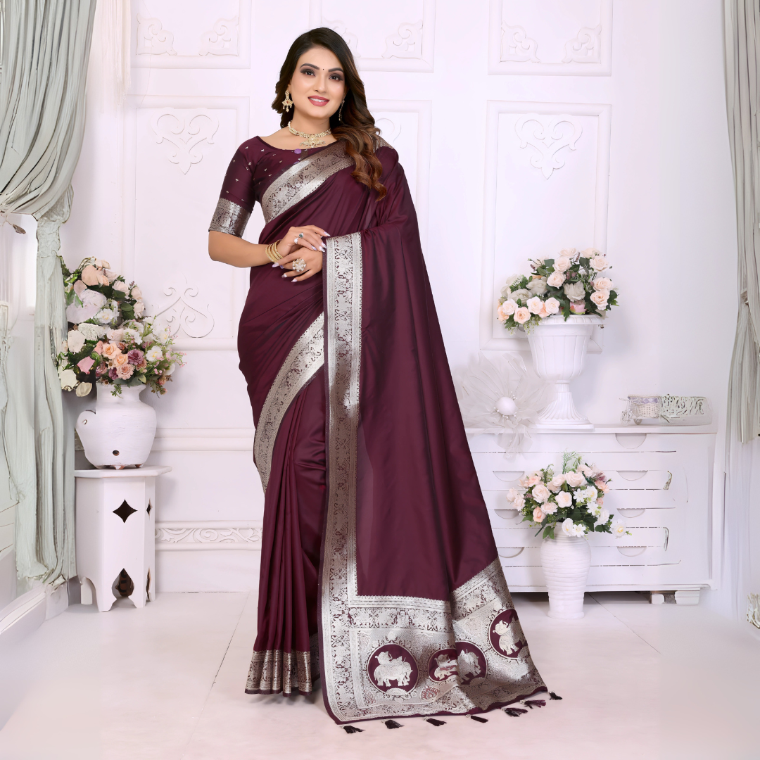 Woman wearing a wine-colored silk saree adorned with an elegant silver zari border and traditional elephant motifs on the pallu, paired with a matching blouse featuring silver detailing. She poses gracefully in a classic white floral-themed room decorated with ornate wall panels and flower arrangements in white vases.