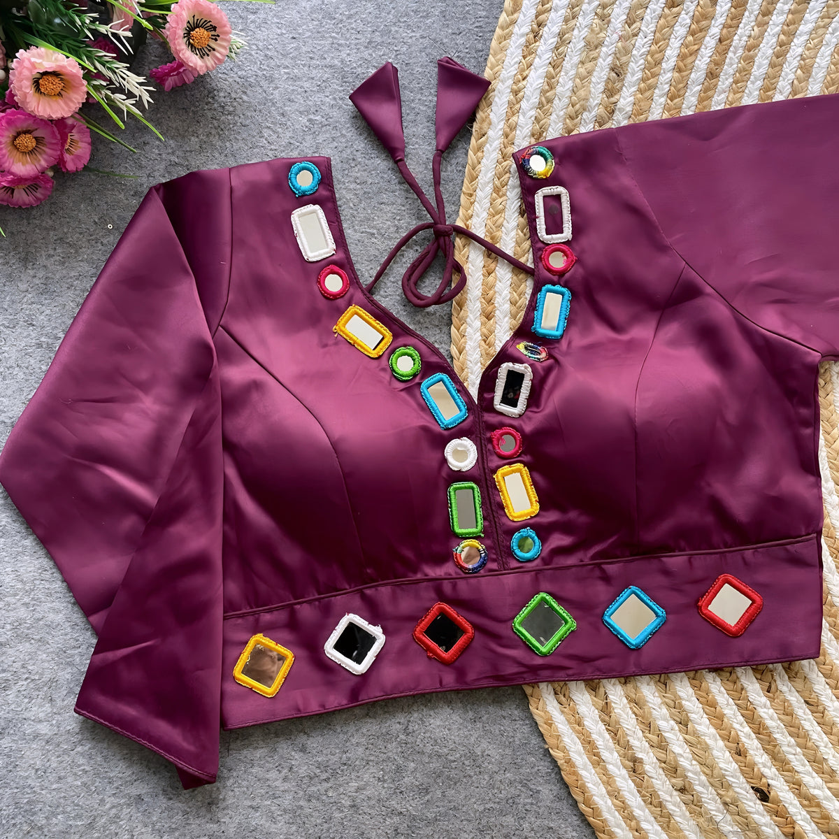 Wine-colored blouse decorated with vibrant square and round mirror work featuring multicolored embroidered borders, stylish back tie-up details, and three-quarter sleeves. Displayed flat on a textured grey surface with a woven mat and pink flowers, highlighting its festive and contemporary ethnic appeal.v