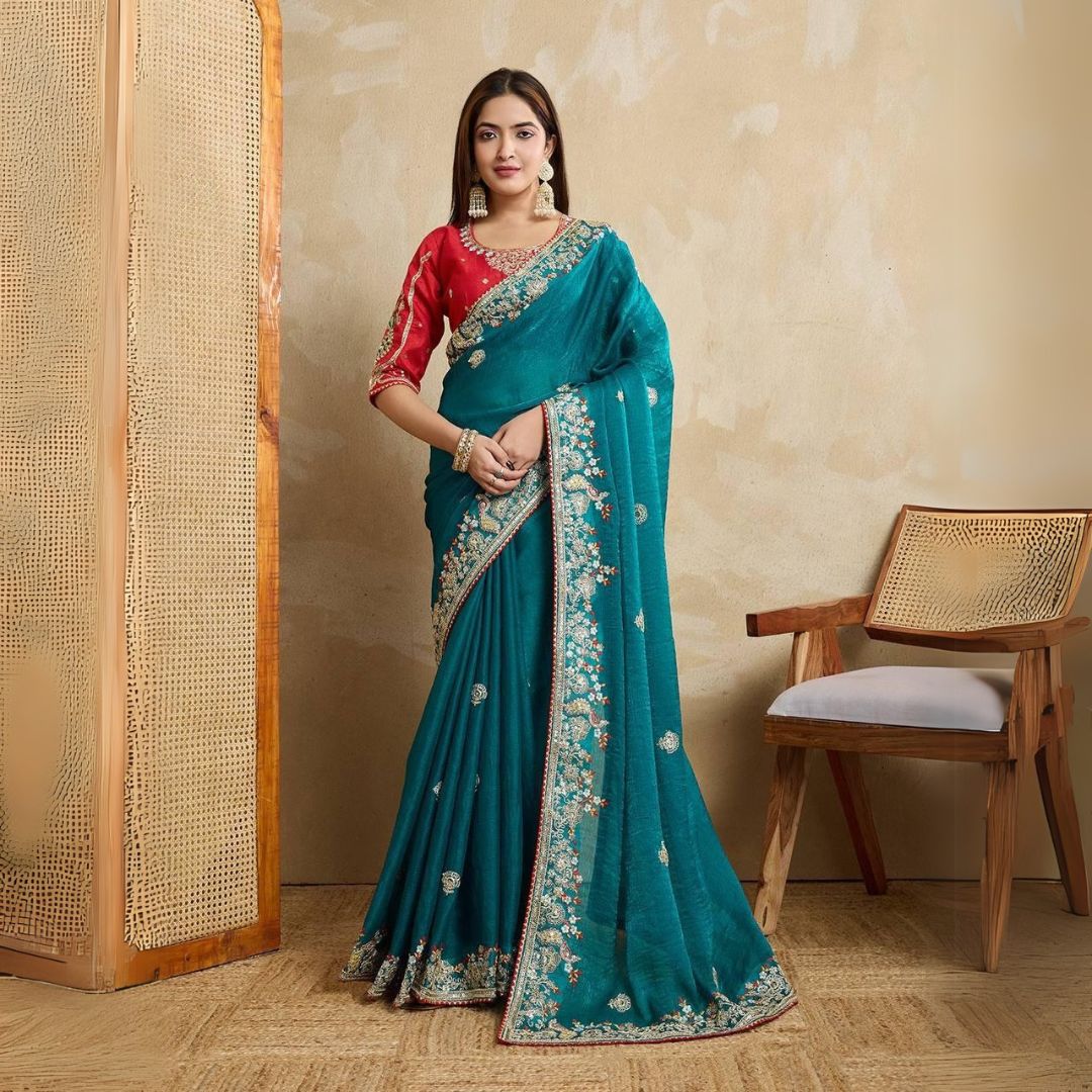 Woman wearing a teal blue embroidered saree with intricate floral border and motifs, paired with a red blouse and traditional jewelry, standing indoors beside a wooden chair and cane screen