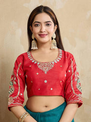 Woman wearing a teal blue embroidered saree with intricate floral border and motifs, paired with a red blouse and traditional jewelry, standing indoors beside a wooden chair and cane screen