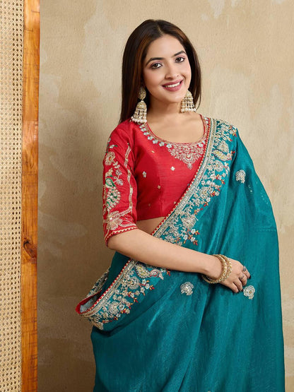 Woman wearing a teal blue embroidered saree with intricate floral border and motifs, paired with a red blouse and traditional jewelry, standing indoors beside a wooden chair and cane screen