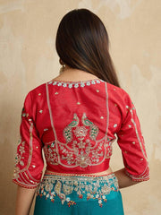 Woman wearing a teal blue embroidered saree with intricate floral border and motifs, paired with a red blouse and traditional jewelry, standing indoors beside a wooden chair and cane screen