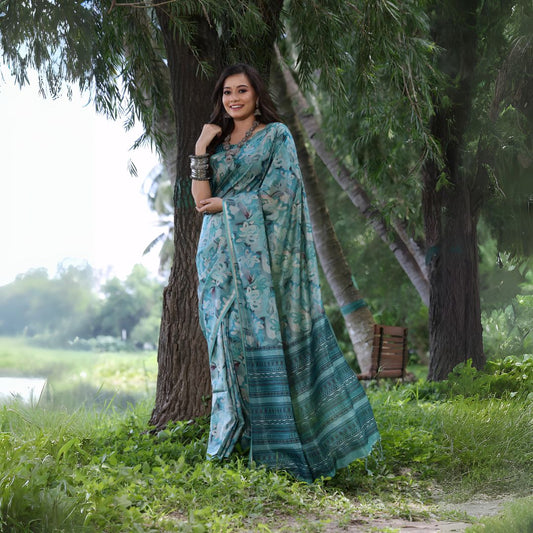 Woman wearing a blue floral silk saree with silver bangles, standing outdoors beside a large tree in a lush green garden.