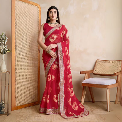 Woman wearing a pink georgette saree with yellow tie-dye patterns and an embellished floral border, paired with traditional jewelry, standing beside a wooden chair and a rattan screen against a beige wall