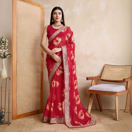 Woman wearing a pink georgette saree with yellow tie-dye patterns and an embellished floral border, paired with traditional jewelry, standing beside a wooden chair and a rattan screen against a beige wall
