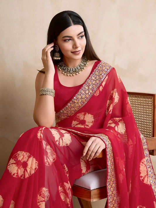 Woman wearing a pink georgette saree with yellow tie-dye patterns and an embellished floral border, paired with traditional jewelry, standing beside a wooden chair and a rattan screen against a beige wall