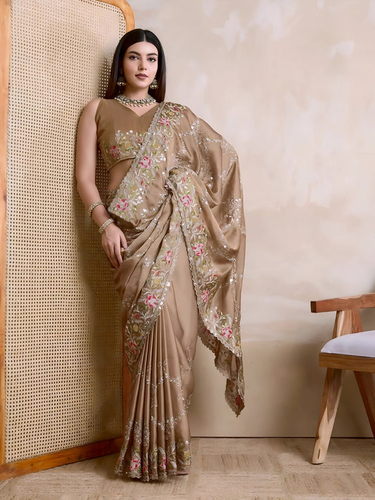 Woman wearing a beige embroidered saree with pink and green floral border, paired with traditional jewelry, standing beside a wooden cane screen and modern chair against a neutral beige wall