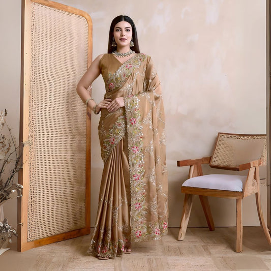 Woman wearing a beige embroidered saree with pink and green floral border, paired with traditional jewelry, standing beside a wooden cane screen and modern chair against a neutral beige wall