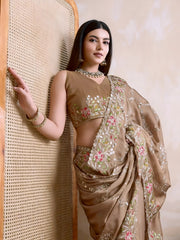 Woman wearing a beige embroidered saree with pink and green floral border, paired with traditional jewelry, standing beside a wooden cane screen and modern chair against a neutral beige wall