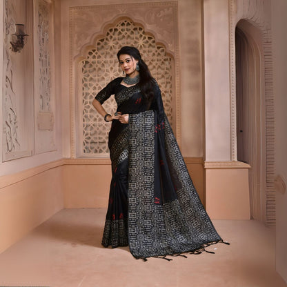 Woman wearing a black silk saree with intricate geometric border and subtle red motifs, paired with traditional silver jewelry, standing in a heritage-inspired room with ornate lattice window and carved walls
