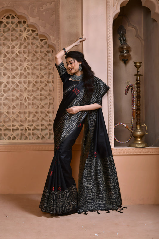 Woman wearing a black silk saree with intricate geometric border and subtle red motifs, paired with traditional silver jewelry, standing in a heritage-inspired room with ornate lattice window and carved walls