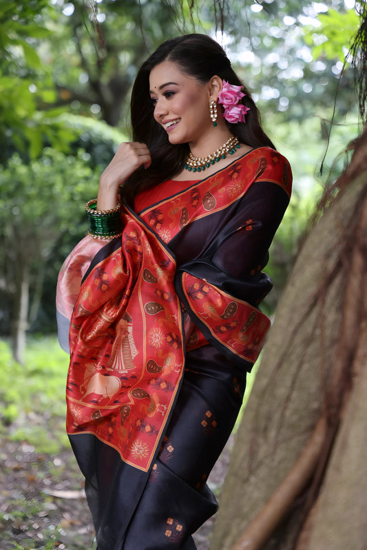 A woman models a striking Black Soft Tussar Silk Saree with small motifs. The saree has a wide, contrasting deep red border and pallu featuring intricate traditional and figurative prints. She wears a long-sleeve red blouse, a green beaded necklace, and is posed in a garden setting.