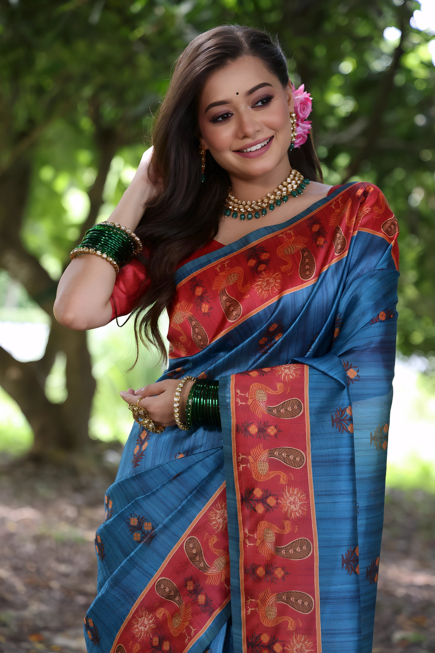 Woman wearing a blue silk saree with a red patterned border and traditional jewelry, standing on a leafy forest path surrounded by lush green trees