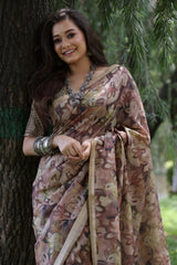 A woman models a Soft Tussar Silk Saree in a muted brown, covered in an all-over artistic print featuring floral motifs. The saree has a wide, woven Zari border with geometric patterns. She wears a complementary blouse and a substantial set of oxidized silver jewelry, posing against a large tree.