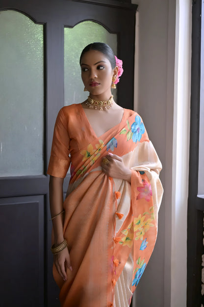 Woman wearing a cream silk saree with pink floral border and pallu, paired with a pink blouse and traditional jewelry, standing indoors near a wooden door with natural light