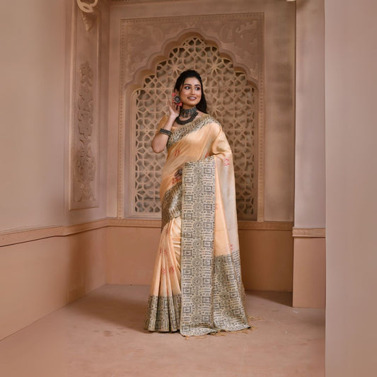 Woman wearing a cream silk saree with a grey geometric border and traditional jewelry, standing in a heritage-inspired room with ornate carved window and pastel walls.