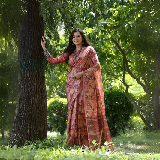  Woman wearing a pink floral silk saree with traditional jewelry and full-sleeve blouse, standing outdoors beside a tree in a lush green garden