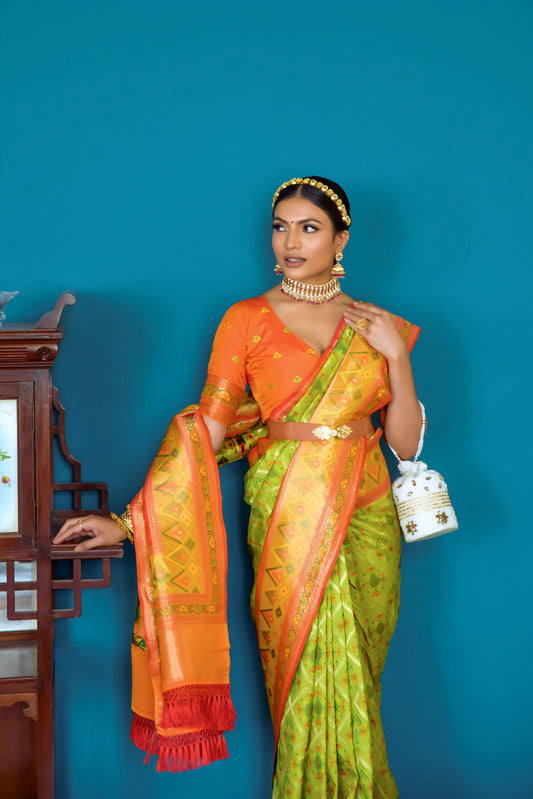 A woman models a striking Lime Green Banarasi Silk Saree with all-over Zari weaving. The wide pallu and border contrast dramatically in Bright Orange with large traditional Zari weaving patterns. She wears an orange blouse, traditional gold jewelry, and a metallic waist belt, posing indoors.