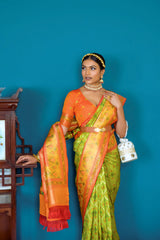 Woman in a green and orange silk saree with gold jewelry standing beside a vintage wooden cabinet in a stylish room with blue walls and marble flooring.