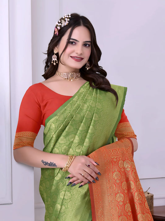 Woman wearing a green silk saree with an orange pallu and red blouse, accessorized with traditional jewelry, standing indoors near decorative plants and a wooden console table