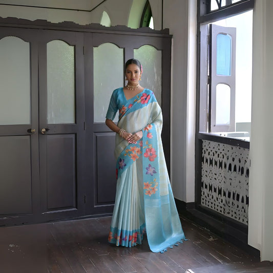 Woman wearing a light blue silk saree with multicolor floral patterns and traditional jewelry, standing indoors near wooden doors and a window with decorative latticework