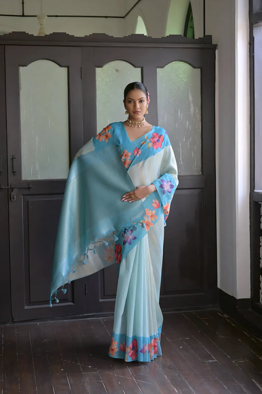 Woman wearing a light blue silk saree with multicolor floral patterns and traditional jewelry, standing indoors near wooden doors and a window with decorative latticework