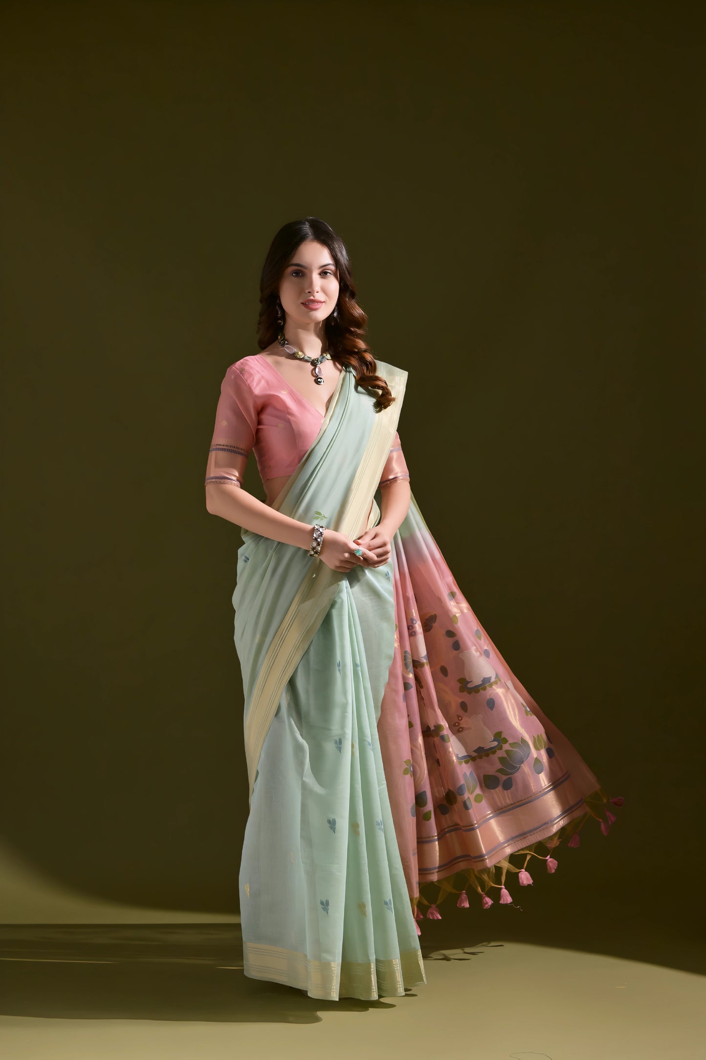 Woman wearing a light green silk saree with a pink floral pallu and tassel border, paired with a pink blouse and silver jewelry, standing against an olive green background