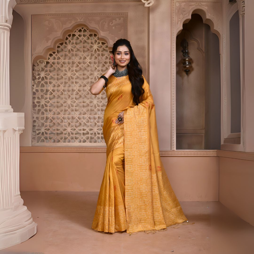 Woman wearing a mustard yellow silk saree with subtle geometric patterns and traditional jewelry, standing in a heritage-inspired room with ornate carved arches and lattice window