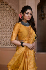 Woman wearing a mustard yellow silk saree with subtle geometric patterns and traditional jewelry, standing in a heritage-inspired room with ornate carved arches and lattice window