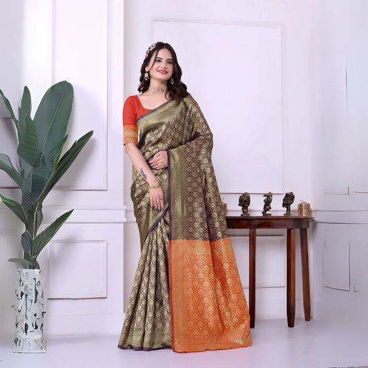 Woman wearing an olive green silk saree with intricate gold motifs and an orange border, paired with a red blouse and traditional jewelry, standing indoors beside a potted plant and wooden console table