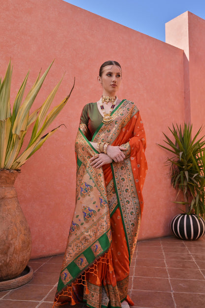 Woman wearing an orange and green silk saree with intricate peacock border and traditional gold jewelry, standing in front of a terracotta wall with potted plants.
