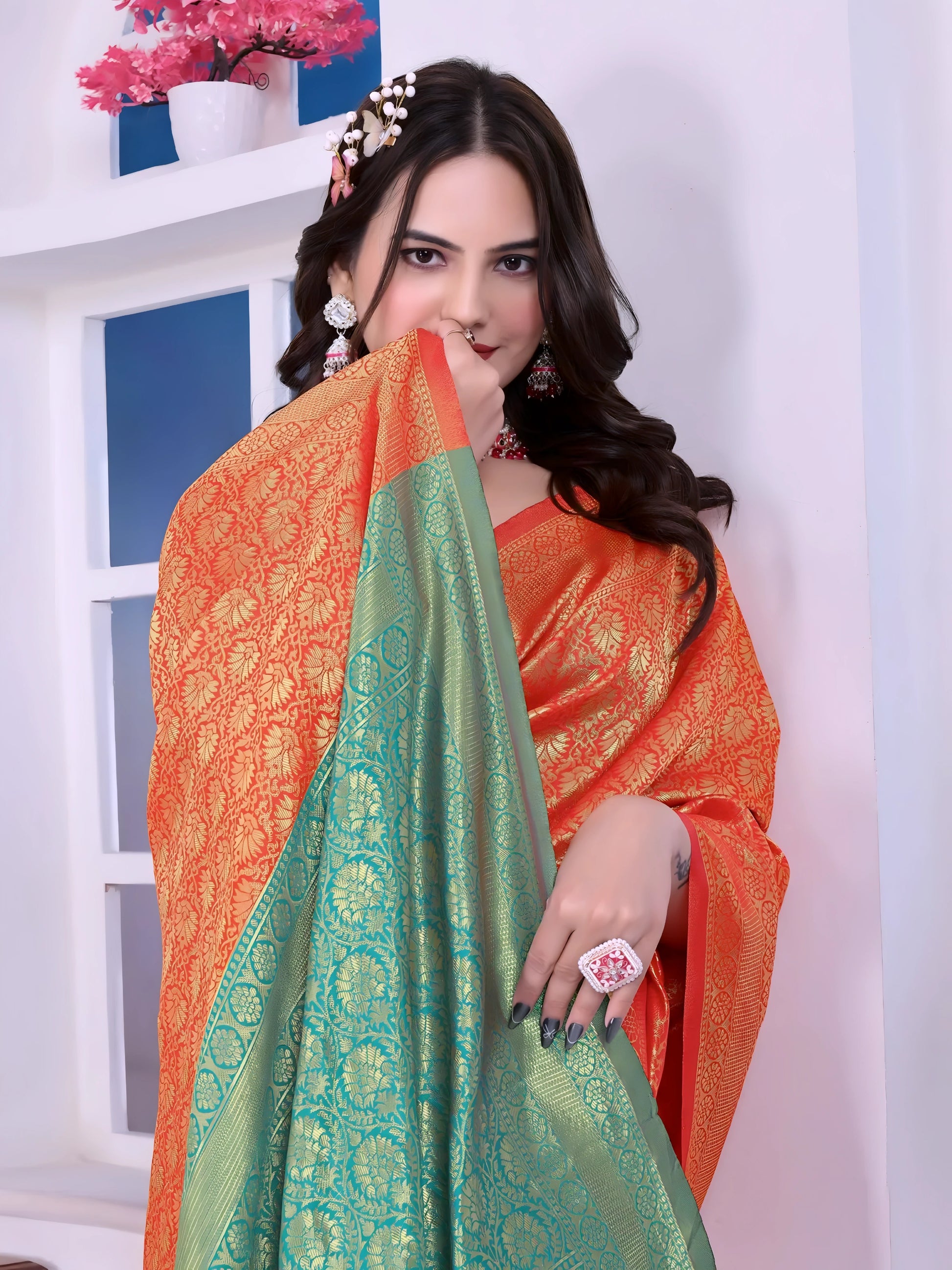 Woman wearing an orange silk saree with intricate golden motifs and a contrasting green pallu, paired with a teal blouse and traditional jewelry, standing indoors near a window and decorative plants.