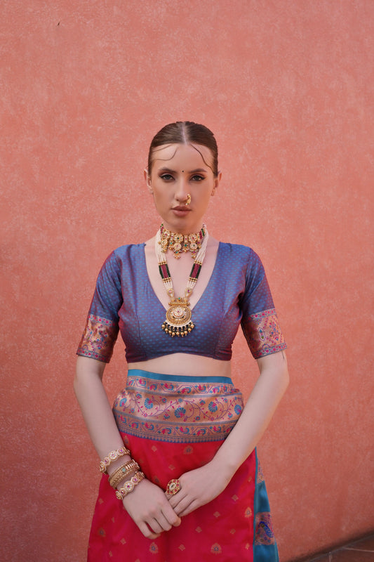 Woman wearing a pink and blue silk saree with intricate patterns and gold jewelry, standing in front of a terracotta wall with potted green plants under a clear blue sky.