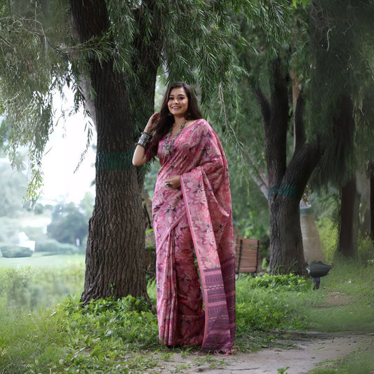 Woman wearing a pink floral silk saree with traditional patterns and silver bangles, standing outdoors beside a large tree in a lush green garden
