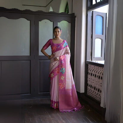 Woman wearing a pink silk saree with colorful floral patterns and a matching blouse, accessorized with traditional jewelry, standing indoors near an open window with natural light

