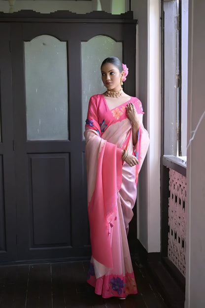  Woman wearing a pink silk saree with multicolor floral patterns and traditional jewelry, standing indoors near a wooden door and window with natural light