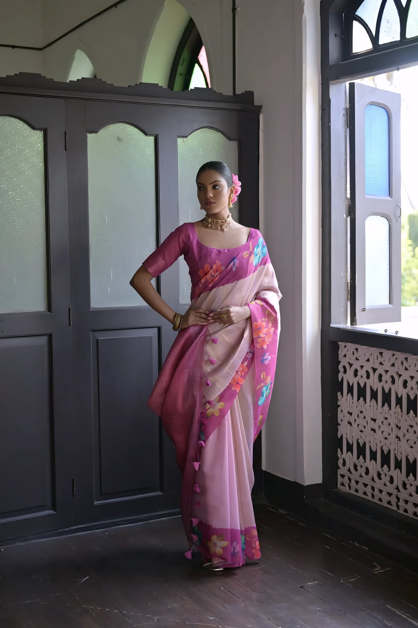 Woman wearing a pink silk saree with colorful floral patterns and a matching blouse, accessorized with traditional jewelry, standing indoors near an open window with natural light.