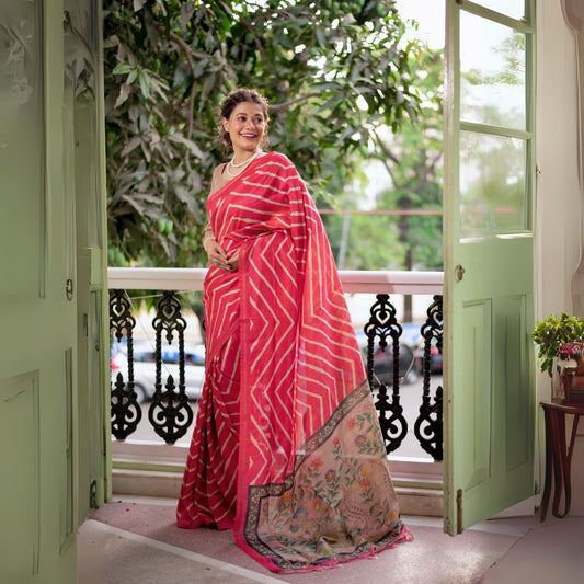 A woman models a vibrant Rose Pink Saree with a bold off-white chevron/striped pattern. The pallu is light beige with a dark border, featuring an elaborate printed motif of colorful flowers and foliage. She wears a pearl necklace and is posed in a doorway with green shutters.