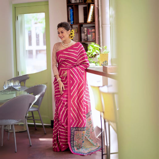 A woman models a vibrant Hot Pink Saree with a bold off-white chevron/striped pattern. The pallu is light beige with a dark border, featuring an elaborate printed motif of colorful flowers. She wears a pearl necklace and a large yellow flower hair accessory, posing indoors.