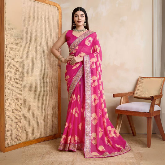 Woman wearing a pink georgette saree with yellow tie-dye patterns and an embellished floral border, paired with traditional jewelry, standing beside a wooden chair against a beige textured wall