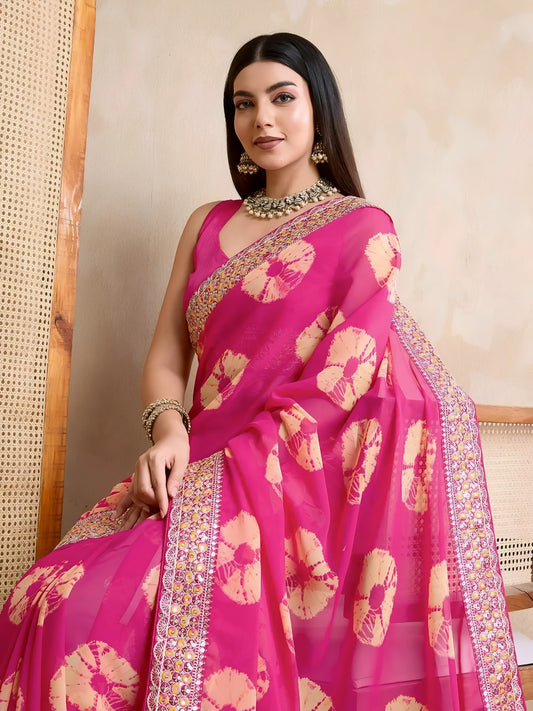 Woman wearing a pink georgette saree with yellow tie-dye patterns and an embellished floral border, paired with traditional jewelry, standing beside a wooden chair against a beige textured wall