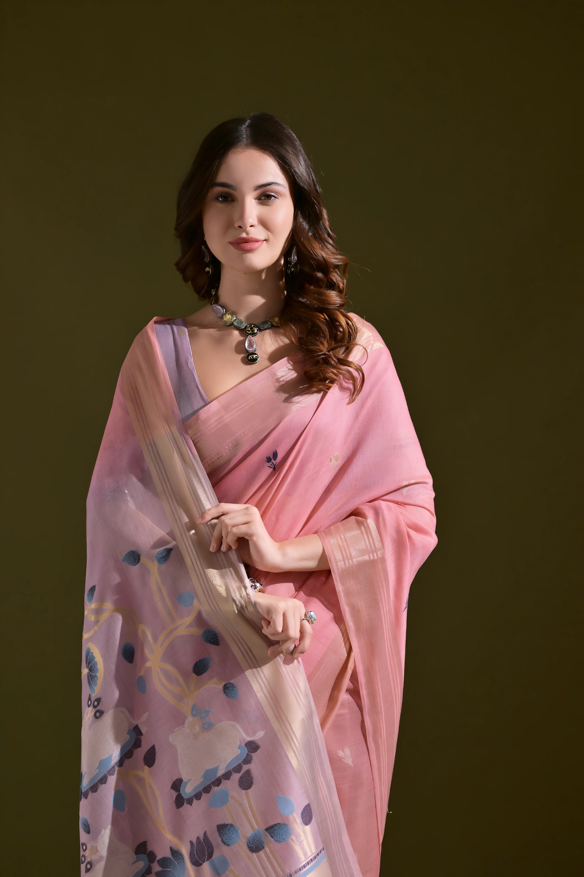 Woman wearing a pink silk saree with a blue floral pallu and tassel border, accessorized with silver jewelry, standing against an olive green background.