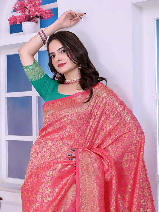 Woman wearing a pink silk saree with golden motifs and a contrasting green pallu, paired with a green blouse and floral hair accessories, posing indoors near a window and wooden console table.