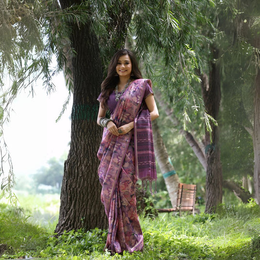 Woman wearing a purple floral silk saree with traditional jewelry, standing outdoors beside a large tree in a lush green garden